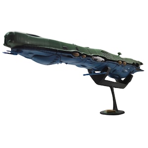 May include: A model spaceship with a green top and blue underside, mounted on a black stand. The spaceship has detailed features, including small protrusions and a textured surface. The stand has a circular base.