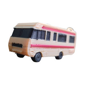 Puede incluir: Una autocaravana beige en miniatura con ventanas y ruedas negras, y una franja roja. La autocaravana de juguete tiene una rejilla delantera detallada y está diseñada para parecerse a un vehículo recreativo.