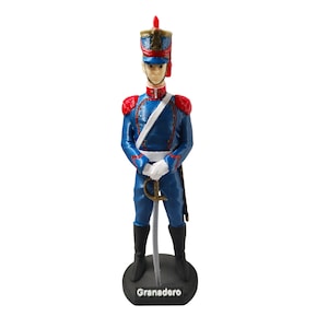 Puede incluir: Figura de un soldado con uniforme azul, charreteras rojas, sombrero alto y espada. La figura está sobre una base negra con la palabra "Granadero". El soldado lleva botas negras y guantes blancos. Aproximadamente 15 cm de altura.