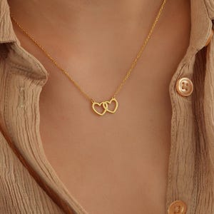 May include: A gold necklace with two interlocking heart pendants. The necklace is worn by a person with a light brown shirt.