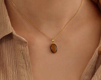 May include: A gold-colored necklace with a pendant featuring an oval, brown gemstone. The necklace is worn against a beige collared shirt. The pendant is suspended from a delicate chain.