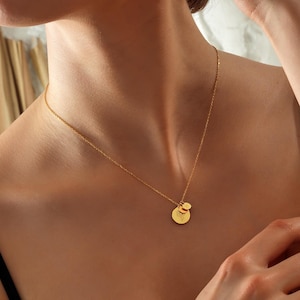 Może przedstawiać: Delikatny złoty naszyjnik z dwoma okrągłymi wisiorkami. Większy wisiorek ma mniejszy, dołączony wisiorek. Naszyjnik jest noszony na ciemnej górze, podkreślając blask złota. Biżuteria to prosty, ale elegancki dodatek.