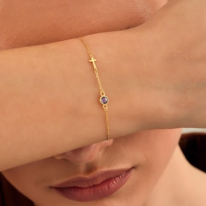May include: A delicate gold bracelet featuring a small cross charm and a purple gemstone. The bracelet is displayed on a wrist, showcasing its fine chain and minimalist design. The jewelry is a subtle accessory.