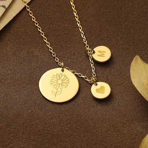 May include: Gold-coloured necklace with three pendants. The largest pendant features a daisy design, while the other two have a letter "M" and a heart. The chain is delicate, and the pendants are circular.