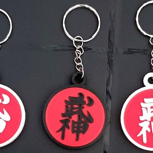 Könnte beinhalten: Drei rote und weiße runde Schlüsselanhänger mit jeweils einem schwarzen Kanji-Zeichen. Das Kanji-Zeichen ist "真神", was "Wahrer Gott" bedeutet.