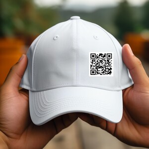 Könnte beinhalten: Eine weiße Baseballkappe mit gebogenem Schirm. Ein quadratischer schwarz-weißer QR-Code ist vorne angebracht. Die Kappe wird von zwei Händen gehalten, mit einem verschwommenen grünen Hintergrund.