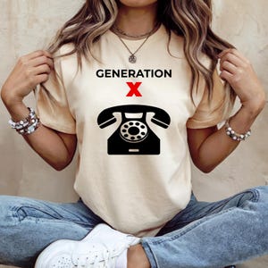 Può includere: T-shirt color crema con la scritta "GENERATION X" sopra una grafica di telefono nero a disco con una "X" rossa al centro. La maglietta è abbinata a jeans blu e scarpe da ginnastica bianche.
