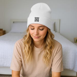 Könnte beinhalten: Eine weiße Mütze mit einem schwarzen QR-Code-Patch. Die Mütze ist aus einem weichen, gestrickten Material gefertigt. Die Mütze wird von einer Person mit blonden Haaren und einem beigefarbenen T-Shirt getragen.