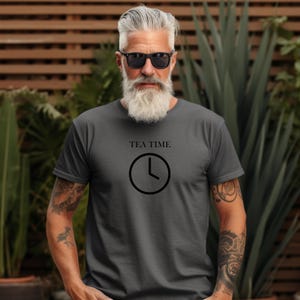 May include: Dark gray t-shirt with the words "TEA TIME" above a clock graphic. The clock shows the time as 4 o'clock. The shirt is short-sleeved and made of a soft material. The person in the image is wearing sunglasses.