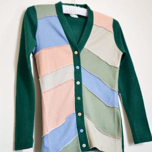 May include: A long-sleeved cardigan in shades of green, blue, pink, and beige hangs on a hanger. The front features a patchwork design with colorful, horizontal stripes. The cardigan has a V-neck and is fastened with colorful buttons.
