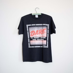 May include: Black t-shirt with the D.A.R.E. logo in red and white, featuring the text "TO RESIST DRIGS AND VIOLENE." The shirt also has the text "DRUG ABUSE RESISTANCE EDUCATION" around the logo.
