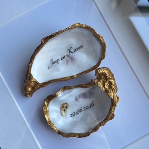 Puede incluir: Dos conchas de ostra con bordes pintados en oro e interiores blancos. Una concha presenta el texto negro "Joep en Karen" y la otra muestra el texto negro "20.06.2026" con dos peque&ntilde;os anillos dorados descansando dentro de la concha.