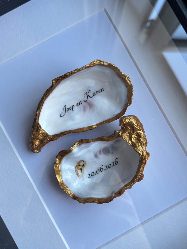 Puede incluir: Dos conchas de ostra con bordes pintados en oro e interiores blancos. Una concha presenta el texto negro "Joep en Karen" y la otra muestra el texto negro "20.06.2026" con dos peque&ntilde;os anillos dorados descansando dentro de la concha.
