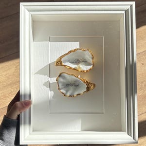 Könnte beinhalten: Ein weißer Schattenfugenrahmen mit zwei Austernschalen, deren Ränder gold bemalt sind. Die obere Schale enthält eine kleine weiße Perle und den Text Sarah & Stephen. Die untere Schale zeigt das in schwarzer Schrift gedruckte Datum 07.09.1996.