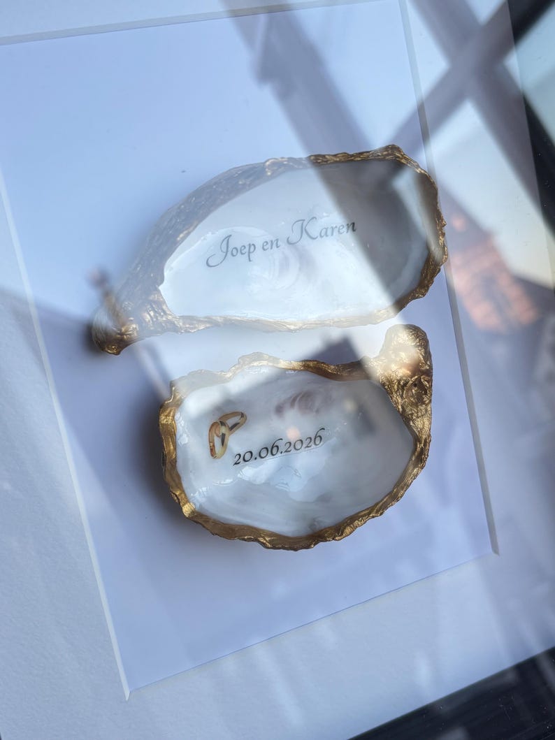Puede incluir: Dos conchas de ostra blancas con bordes pintados en dorado dentro de un marco. La concha superior presenta el texto Joep en Karen en letra cursiva negra, mientras que la inferior muestra la fecha 20.06.2026 y dos peque&ntilde;os anillos dorados.