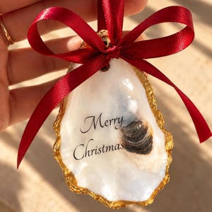 Könnte beinhalten: Ein dekoratives Austernschalen-Ornament mit einer glatten, perlmuttweißen Oberfläche und dem schwarzen Schriftzug Merry Christmas auf der Vorderseite. Die Ränder der Schale sind mit metallischem Gold verziert und ein dunkelrotes Satinband ist oben zu einer Schleife gebunden.