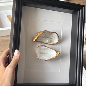 May include: A black shadow box frame containing two oyster shells with gold-painted edges. The top shell features the text "Lucy and Michael" and a white pearl. The bottom shell features the date "15.08.2026" printed in black on the white interior.