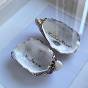 Puede incluir: Dos conchas de ostra naturales con interiores lisos y nacarados. Una concha tiene inscrita la frase Michelle & Alessandro en letra cursiva negra, y la otra concha muestra la fecha 20. Juni 2026 en letras negras a juego.
