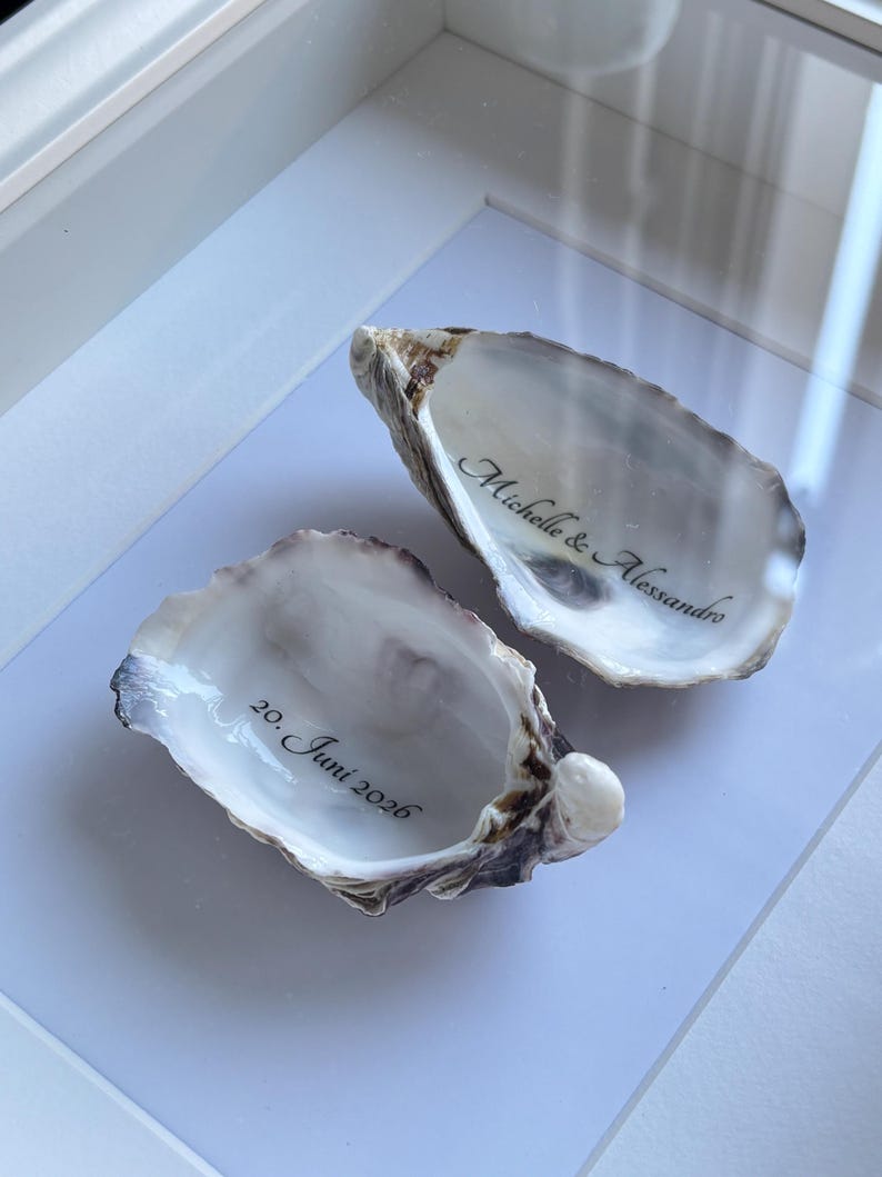 Puede incluir: Dos conchas de ostra naturales con interiores lisos y nacarados. Una concha tiene inscrita la frase Michelle & Alessandro en letra cursiva negra, y la otra concha muestra la fecha 20. Juni 2026 en letras negras a juego.