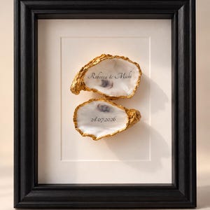 Pode incluir: Uma moldura retangular preta contendo duas conchas de ostra brancas com bordas pintadas de dourado. A concha superior apresenta o texto Rebecca & Michi, e a inferior a data 24.07.2026. As conchas estão montadas num passe-partout branco.