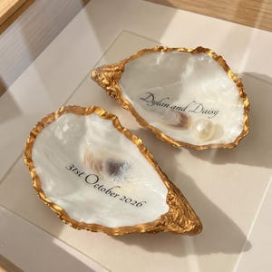 Op de afbeelding: Twee witte oesterschelpen met goudkleurige randen. Eén schelp bevat de tekst "Dylan and Daisy" en een kleine witte parel. De andere schelp toont de tekst "31st October 2026" in zwarte letters. Beide schelpen hebben een gladde, parelmoerachtige binnenkant.