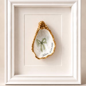 May include: A white framed wall art piece featuring a single oyster shell with a gold-painted edge. The interior of the shell displays a painted sage green ribbon bow and the black text Julian 17-09-2026 centred on a white mat board.