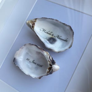 Puede incluir: Dos conchas de ostra naturales expuestas dentro de un marco blanco. Una concha presenta el texto impreso en negro Michelle & Alessandro, mientras que la otra muestra el texto impreso en negro 20. Juni 2026. Las conchas tienen interiores nacarados.