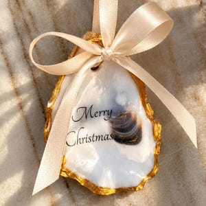Könnte beinhalten: Ein dekoratives Austernschalen-Ornament mit einer glatten weißen Innenseite und dem schwarzen Schriftzug Merry Christmas auf der Oberfläche. Die Schale hat einen goldfarbenen Rand und ist oben mit einer hellbeigen Satinschleife versehen.