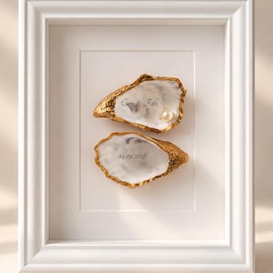 Könnte beinhalten: Ein weiß gerahmtes Wandbild mit zwei Austernschalen mit goldfarbenen Rändern. Die obere Schale ist mit dem Text "Caro & Fabi" beschriftet und enthält eine Perle. Die untere Schale ist mit dem Datum "04.09.2026" versehen.