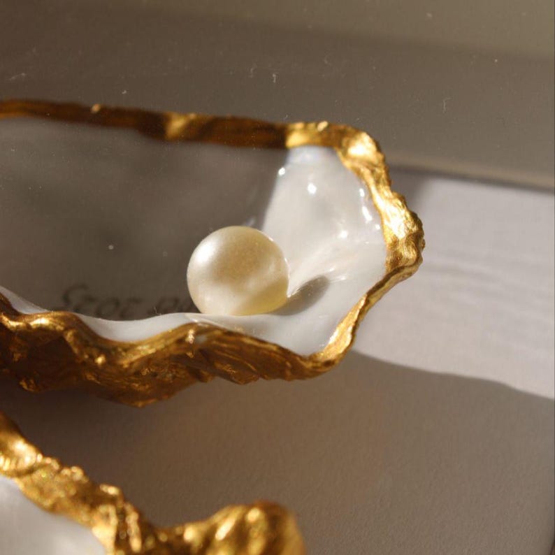 Puede incluir: Una perla blanca, lisa y redondeada descansa dentro de una concha de ostra decorativa. La concha presenta un interior blanco brillante y un borde texturizado pintado en oro met&aacute;lico. La forma es curva, org&aacute;nica y con bordes irregulares.