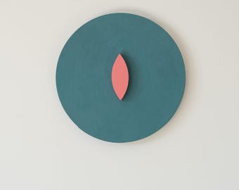 May include: A teal-colored, circular wall art piece with a salmon-colored, leaf-shaped accent in the center. The artwork is mounted on a white wall, creating a minimalist and modern aesthetic. The piece is a simple, yet striking, geometric design.