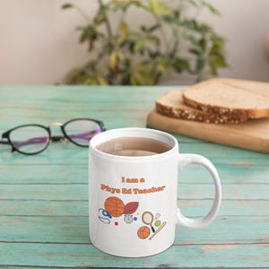 Puede incluir: Taza de cerámica blanca con el texto "I am a Phys Ed Teacher" en naranja y gráficos deportivos. La taza contiene un líquido marrón claro y está sobre una superficie de madera turquesa.