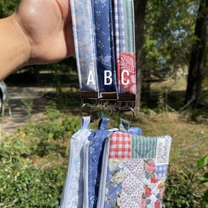 May include: A hand holding four fabric wristlet keychains. The keychains feature various blue and white floral and patchwork patterns. Each keychain has a metal ring and a zipper pouch. The text "Oh Sew Darling" is visible on one pouch.