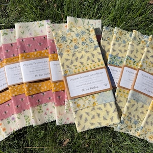 May include: A collection of organic beeswax food wraps featuring various floral and bee patterns in yellow, pink, and blue. Each wrap is secured with a paper band labeled Organic Beeswax Food Wraps Starter Pack 18x8, 10x10 by Oh Sew Darling.