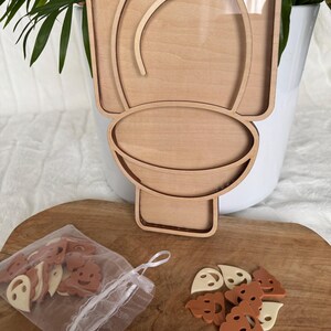 Peut inclure: Un cadre en bois clair en forme de toilettes avec une façade en acrylique transparent. À côté se trouve un petit sac à cordon en organza blanc contenant plusieurs petits jetons en plastique plats, marron et crème, en forme d'émoticônes.