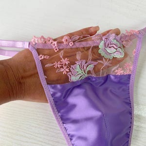 Puede incluir: Tanga de satén lila con bordado floral. El tanga presenta un panel de malla transparente con flores bordadas en rosa y verde y un delicado ribete de encaje rosa. La tela de satén tiene un brillo sutil.
