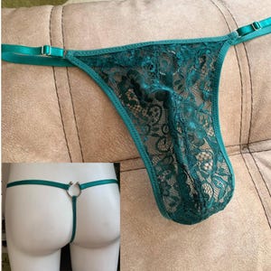 Könnte beinhalten: Ein dunkel smaragdgrüner Spitzen-Tanga mit Blumenmuster, verstellbaren Satinbändern mit silberfarbenen Metallschiebern und einem mittigen silberfarbenen Metallring am Rückenteil, der die dünnen elastischen Bänder miteinander verbindet.