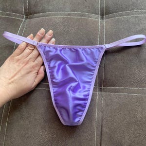 Op de afbeelding: Een lavendelkleurige string met een gladde, satijnachtige afwerking. Het kledingstuk is voorzien van dunne, bijpassende stoffen zijbandjes en een driehoekig voorpand met gestikte randen, wat zorgt voor een strak en eenvoudig ontwerp voor dit ondergoed.