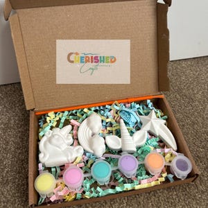 K&ouml;nnte beinhalten: Ein Bastelset in einem Karton, mit wei&szlig;en Gips-Einhornfiguren, einem Stern und Farbt&ouml;pfen in verschiedenen Farben. Die Box ist mit bunten Papierschnipseln gef&uuml;llt und tr&auml;gt den Text "Cherished Crafts" auf dem Deckel.
