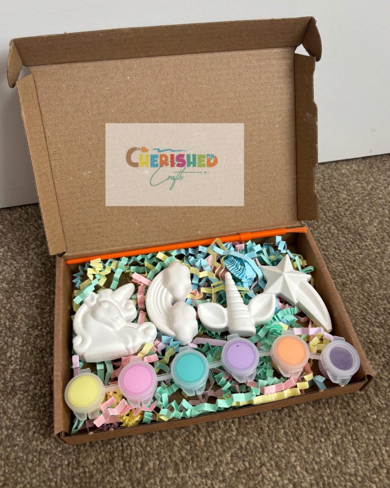 K&ouml;nnte beinhalten: Ein Bastelset in einem Karton, mit wei&szlig;en Gips-Einhornfiguren, einem Stern und Farbt&ouml;pfen in verschiedenen Farben. Die Box ist mit bunten Papierschnipseln gef&uuml;llt und tr&auml;gt den Text "Cherished Crafts" auf dem Deckel.
