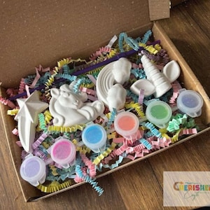K&ouml;nnte beinhalten: Ein Bastelset in einem Karton mit wei&szlig;en Einhorn-, Stern- und Regenbogenfiguren. Das Set enth&auml;lt kleine Farbt&ouml;pfe in verschiedenen Farben und bunte Papierstreifen. Die Box ist ge&ouml;ffnet und zeigt den Inhalt.