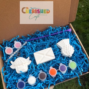 May include: A craft kit in a cardboard box filled with blue paper shreds. It includes a white highland cow figure, a white envelope, a white thistle, a purple paintbrush, a strip of six paint pots, and a strip of two paint pots with rhinestones.