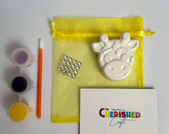 Könnte beinhalten: Ein Bastelset mit einer weißen, giraffe-förmigen Gipsfigur, drei Farbtöpfen, einem Pinsel und einem kleinen Beutel mit Strasssteinen. Das Set enthält einen gelben Kordelzugbeutel und eine Karte mit dem Text "CHERISHED Crafts".