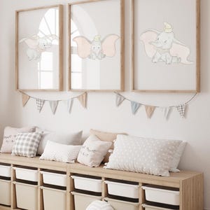 May include: Three framed prints featuring Dumbo the elephant in various poses, with soft pink and gray tones. The prints are displayed above a wooden storage bench with white bins and decorative pillows, creating a nursery or children's room theme.