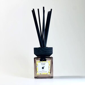 May include: A square glass reed diffuser with a dark wooden top and black reeds. The label reads "GOLDEN ELIXIR" and "NO. 4 REED DIFFUSER" with a lemon illustration. The bottle is filled with a light-colored liquid.