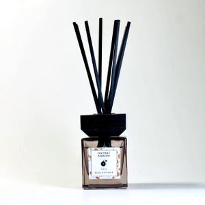 May include: A square glass reed diffuser with black reeds. The label reads "Coconut Paradise" and "No. 4 Reed Diffuser". The diffuser has a dark wooden top and is filled with a light brown liquid. The reeds are black and stand upright.