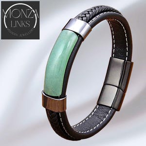 May include: Black leather bracelet with a braided top section and a light green stone accent. The bracelet has a silver clasp and is stitched with white thread. The text "MONZA LINKS MODERN JEWELLERY" is in the upper left corner.