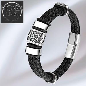 以下が含まれることがあります： 黒い編み込みレザーブレスレット、シルバーカラーのメタルアクセント付き。 ブレスレットは、幾何学模様の中央装飾部分と、円形のロゴに「MONZA LINKS MODERN JEWELLERY」のテキストが特徴です。