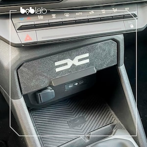 Könnte beinhalten: Schwarze Autokonsole mit silbernem "DC"-Logo. Die Konsole verfügt über einen USB-Anschluss und einen Ladeanschluss. Sie befindet sich zentral im Armaturenbrett, unterhalb der Klimabedienelemente.