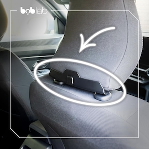 Könnte beinhalten: Ein schwarzer Kunststoffhaken als Zubehör für Autokopfstützen mit geschwungenem Design, montiert an der Basis einer grauen Stoffkopfstütze. Das Produkt verfügt über einen zentralen Clip und zwei abgerundete Stützfüße, Markenname boblab sichtbar.